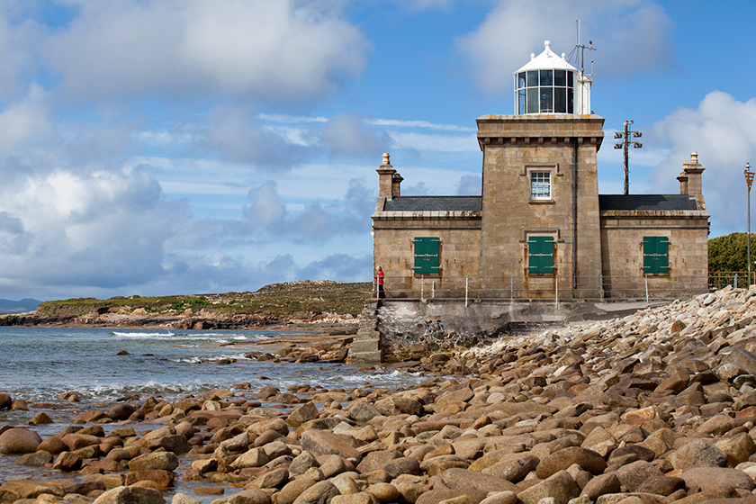 El faro de Blacksod, de piedra cuadrada y con persianas verdes, se encuentra frente a una costa rocosa bajo nubes y una marea tranquila.