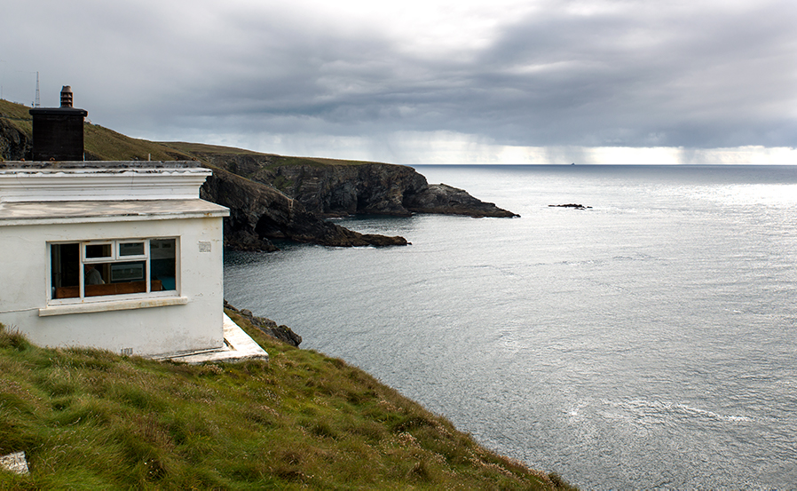 Una baja estación de señalización blanca en el extremo de un acantilado cubierto de hierba en Mizen Head con vistas al mar tranquilo bajo un cielo gris y lluvioso.