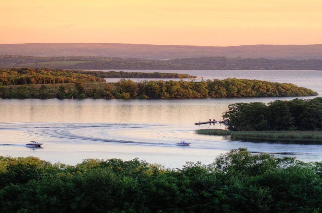 Motorboats leaving curved wakes on Lough Erne at sunset with small forested islands in view.