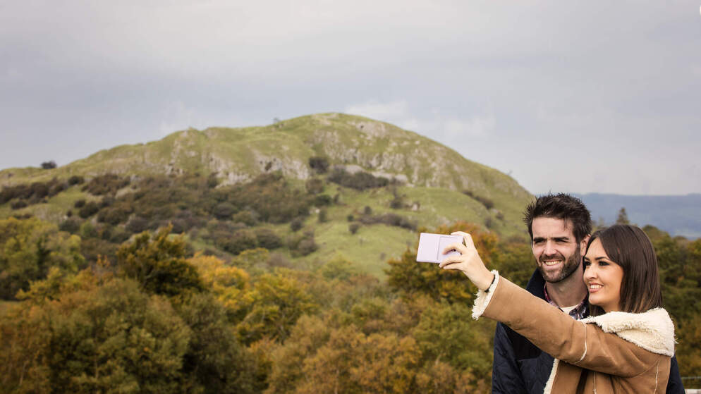 Couple taking a selfie against a lush green hillside in Ireland under a soft grey sky.