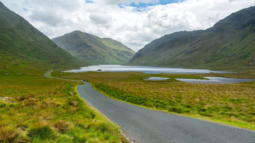 View of winding road through green mountains in Connemara, County Galway under cloudy sky.