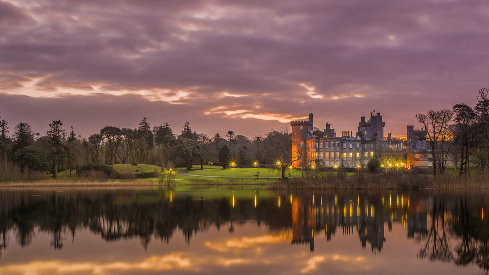 Majestic lakeside Dromoland Castle in County Clare, lit with golden lights against a purple and orange sunset sky.
