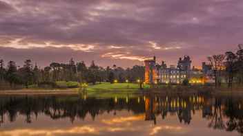 Majestic lakeside Dromoland Castle in County Clare, lit with golden lights against a purple and orange sunset sky.