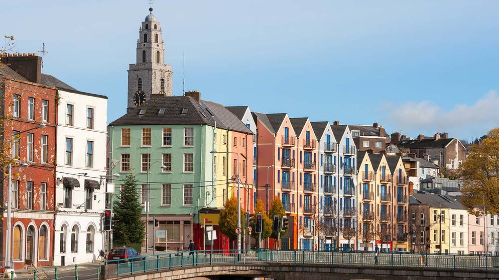 Bunte Häuser entlang einer Straße in Cork mit dem Shandon-Turm im Hintergrund.