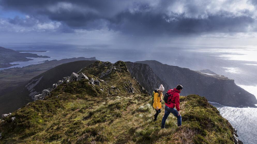 Walkers in bright jackets hiking along a high coastal ridge overlooking sea under heavy clouds.