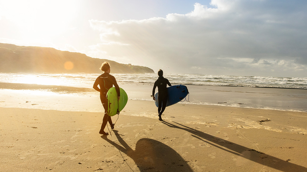 Two surfers carrying boards along Bundoran Beach, County Donegal, at sunrise with waves crashing in.