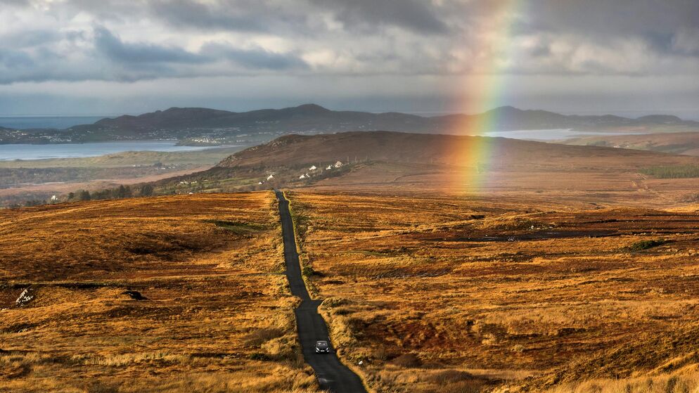 Un arcobaleno si estende su un paesaggio drammatico, con una strada che si snoda tra colline marroni verso montagne e mare lontani.