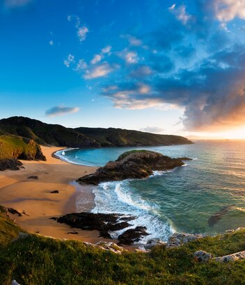 Blick auf den Murder Hole Beach im County Donegal zur goldenen Stunde, mit Klippen, türkisfarbenem Meer und sonnenbeschienenem Atlantikhorizont.