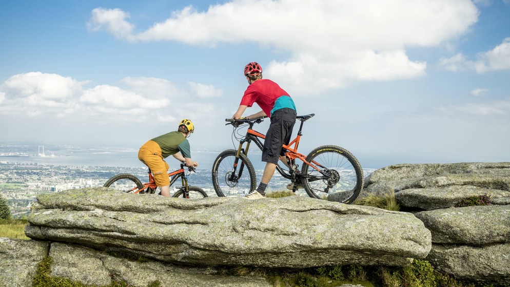 Zwei Radfahrer mit Helmen, die auf Mountainbikes sitzen, machen eine Pause auf einem Felsvorsprung mit Blick auf Dublin und die Bucht von Dublin in der Ferne.