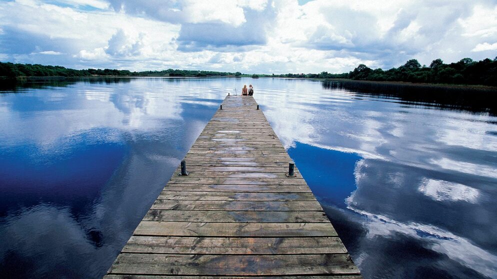 Due persone sedute alla fine di un molo di legno su un Lough Erne calmo a Fermanagh, sotto nuvole drammatiche riflesse nell'acqua.