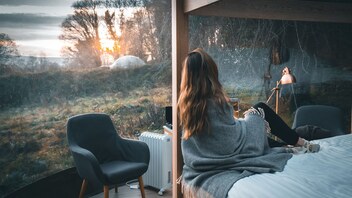 Woman wrapped in a blanket gazing out at woodland through glass walls at Finn Lough, County Fermanagh.