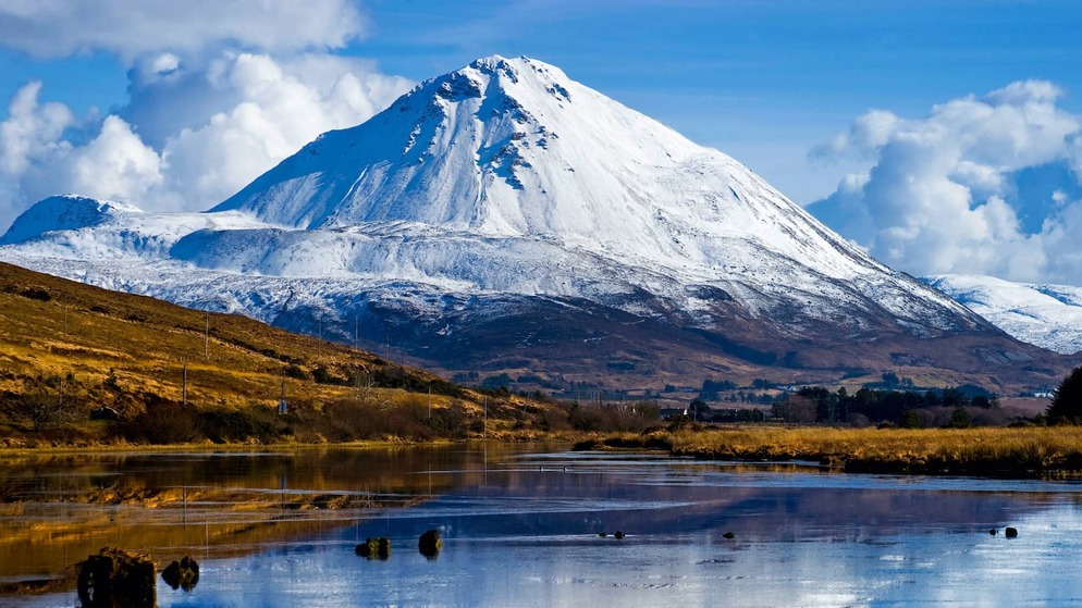 Snow-capped Errigal in County Donegal reflected in a still lake under bright blue sky.