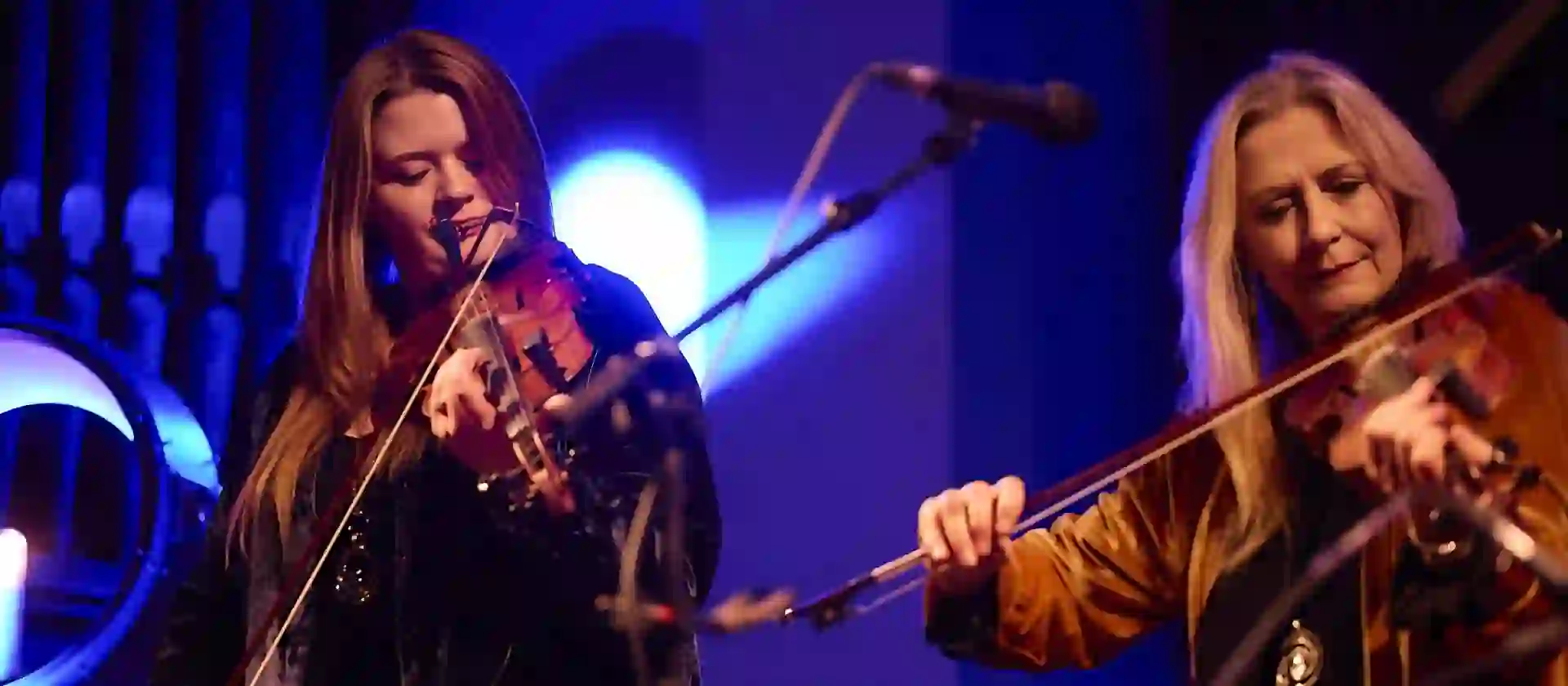 Two musicians from Altan play fiddles on stage under blue lighting during Tradfest, a live traditional Irish music festival in Dublin.