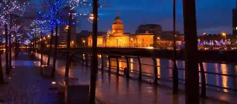 Blue dusk light reflecting on the Liffey beside the glowing Custom House, with tree-lined paths lit by cool, twinkling lights.
