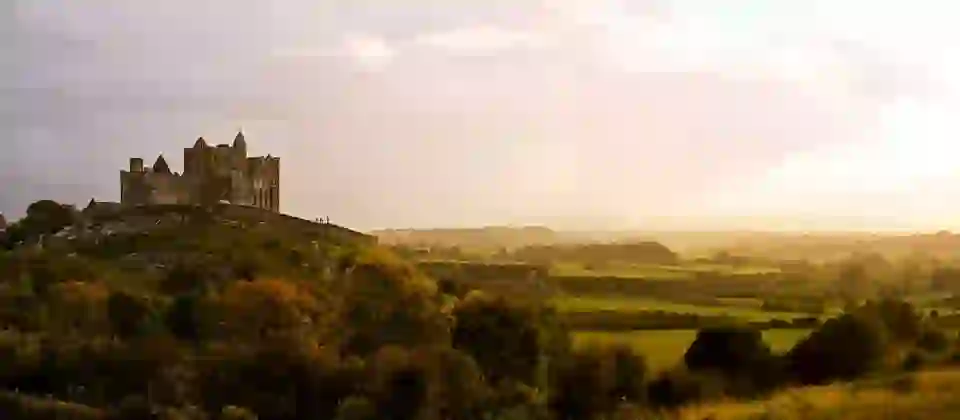 Soft golden light over the Rock of Cashel rising above rolling green and autumn-tinged fields under a hazy sky.