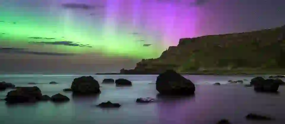 Green and purple aurora shimmering above the Causeway Coast, with dark rocks and calm water in the foreground.
