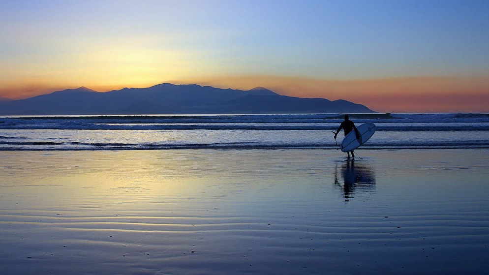 Surfer carrying a board walks at sunset along a beach on the west coast of Ireland.