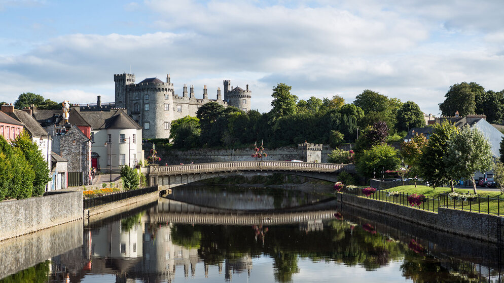 Pont en pierre au-dessus de la rivière Nore face au château de Kilkenny et aux bâtiments de la vieille ville entourés d'arbres et de fleurs.