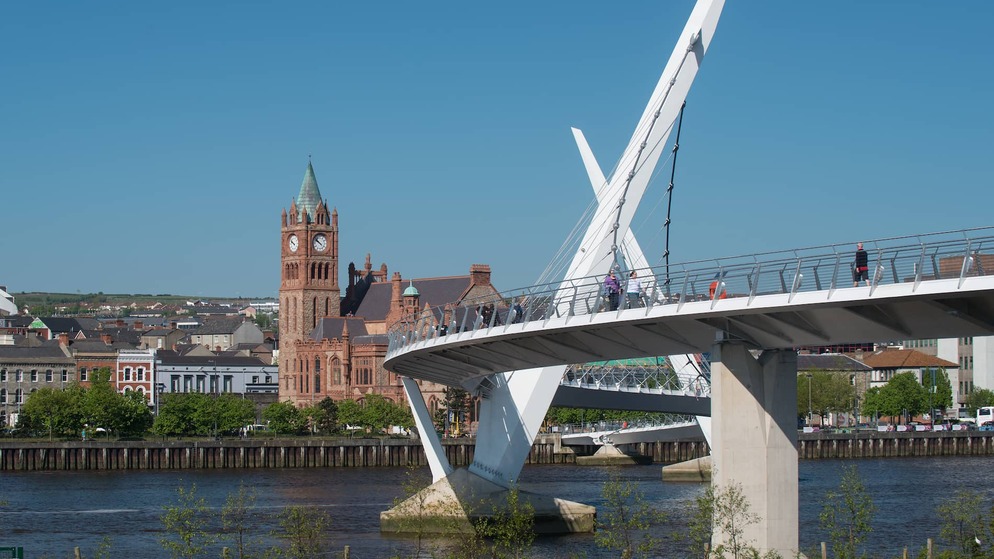 El Puente de la Paz cruza el río Foyle con gente caminando y la Guildhall de Derry de ladrillo rojo al fondo.
