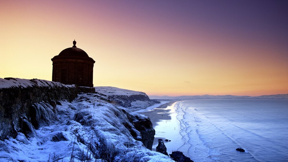 Le scogliere coperte di neve portano al Tempio di Mussenden, che si staglia contro un tramonto invernale dorato su Downhill Beach.