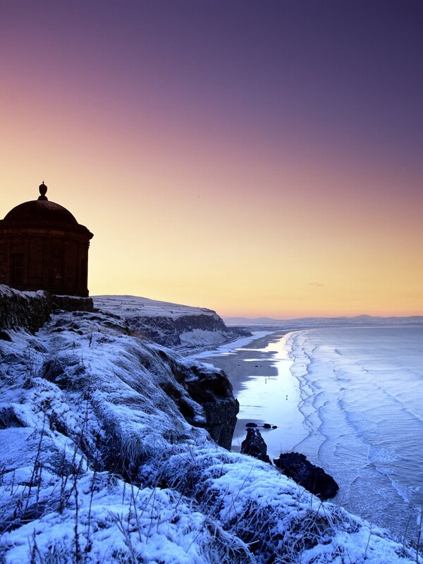 Los acantilados cubiertos de nieve conducen al Templo de Mussenden, siluetado contra un atardecer dorado de invierno sobre la playa de Downhill.