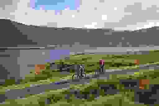 Due ciclisti percorrono un sentiero sul lago di Mayo, con le dorate brughiere e le pendici delle montagne che si riflettono nelle acque ferme.