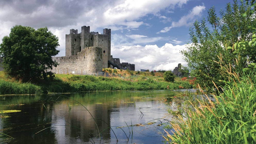 Trim Castle stands by the River Boyne in County Meath, surrounded by green fields and a partly cloudy sky.