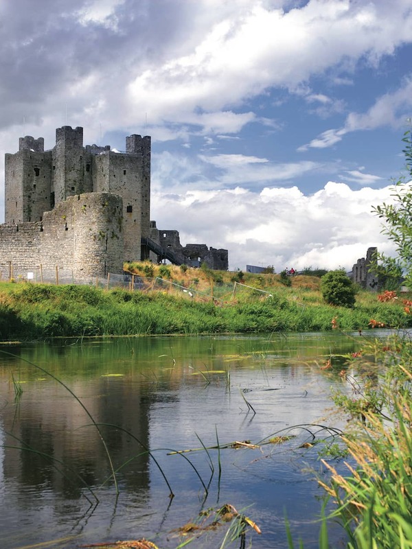 Trim Castle stands by the River Boyne in County Meath, surrounded by green fields and a partly cloudy sky.