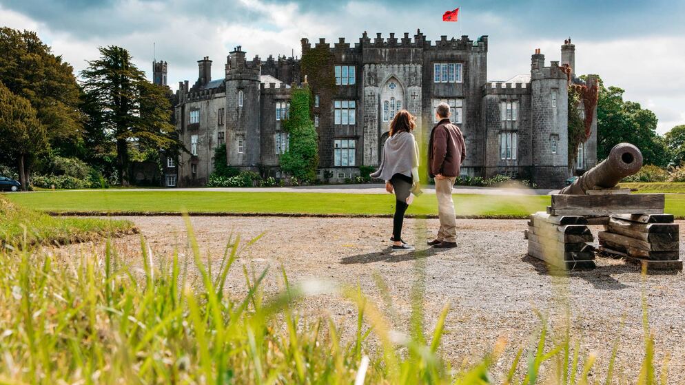 Couple walking towards Birr Castle, a historic grey stone building with turrets and a red flag, County Offaly.