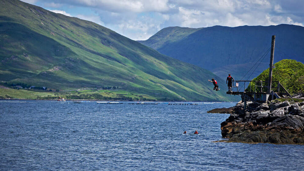 Twee mensen springen van een houten platform in de Killary Fjord, met steile groene bergen op de achtergrond in County Galway.