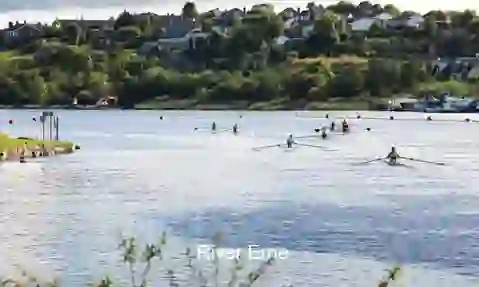 Rowers glide across the River Erne with lush greenery and riverside houses in the background.