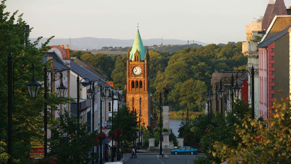 The red-brick Guildhall clock tower rising above a leafy street in Derry~Londonderry on a clear summer evening.