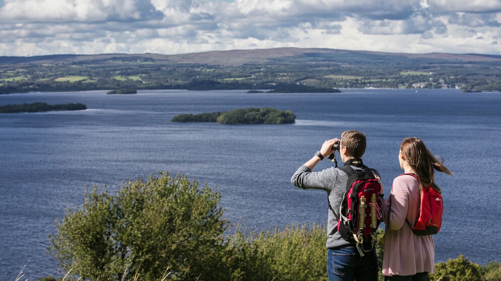 Zwei Wanderer mit Rucksäcken blicken über Lough Derg in der Grafschaft Tipperary, einer betrachtet die Szene durch ein Fernglas.
