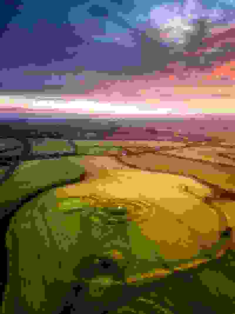 Luchtfoto van de Hill of Tara bij zonsopgang, met patchwork-velden die gloeien onder een roze en oranje hemel.
