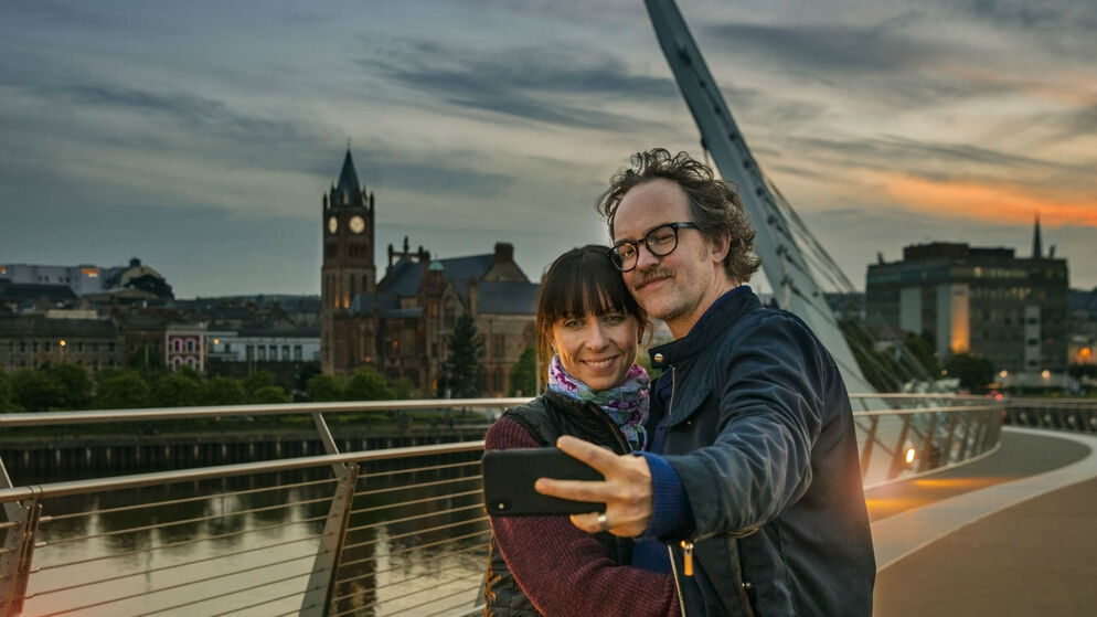 Die Friedensbrücke und das Rathaus in Derry~Londonderry an einem strahlenden Tag mit der Stadtkulisse im Hintergrund.