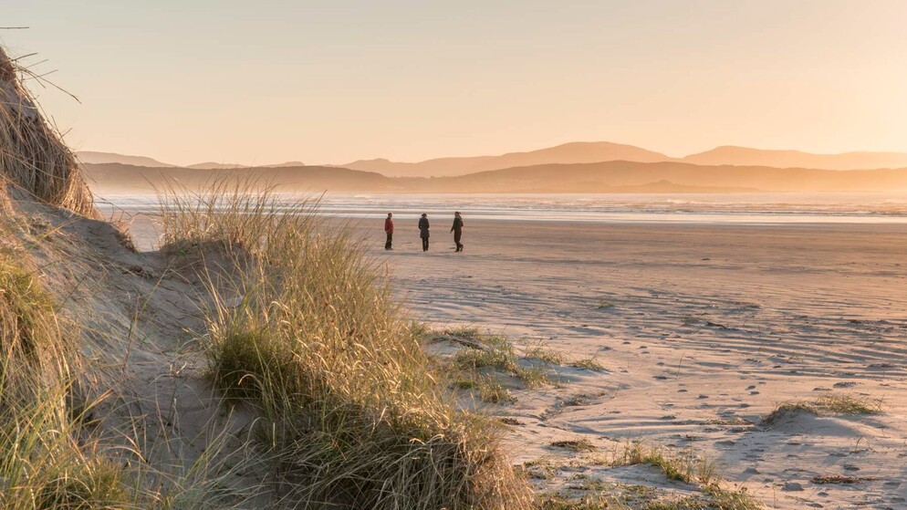 Drei Wanderer überqueren Dooey Beach in der Grafschaft Donegal bei Sonnenuntergang, mit Sanddünen und nebligen Bergen im Hintergrund.