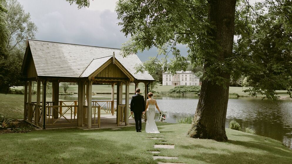 ride and groom walk hand in hand toward a lakeside gazebo, with a Georgian mansion in the background surrounded by greenery.