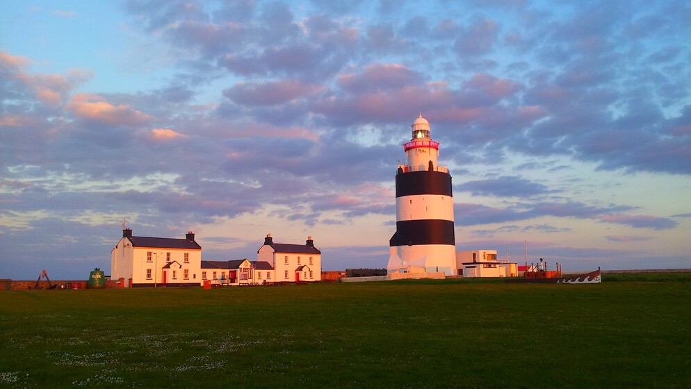 Le phare de Hook avec ses audacieuses bandes noires et blanches au coucher du soleil, à côté de cottages blancs sous un ciel côtier rose-violet.