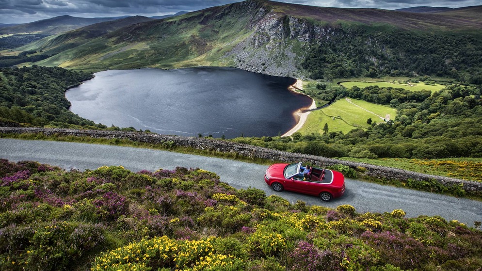 Red convertible car driving along a lakeside road in Wicklow’s mountainous landscape on Ireland’s Ancient East.