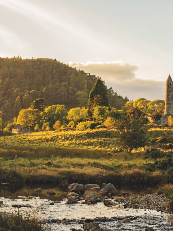 Torre rotonda e rovine monastiche circondate da una luce dorata e da un bosco a Glendalough, nella contea di Wicklow.