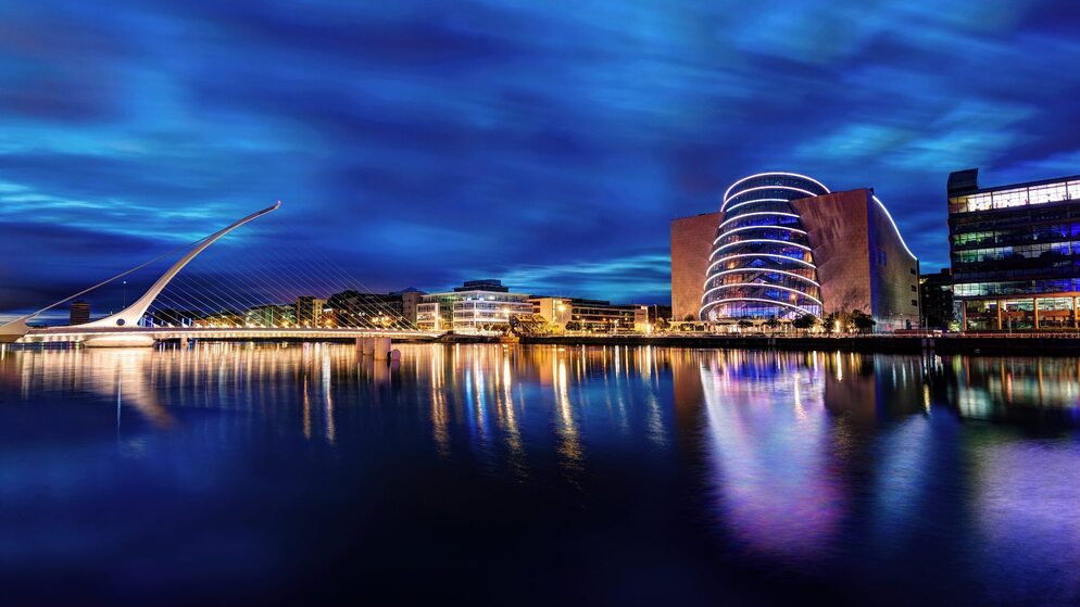 Nachtzicht van Samuel Beckett Bridge met het Convention Centre verlicht in regenboogkleuren die weerspiegelen over de rivier de Liffey.