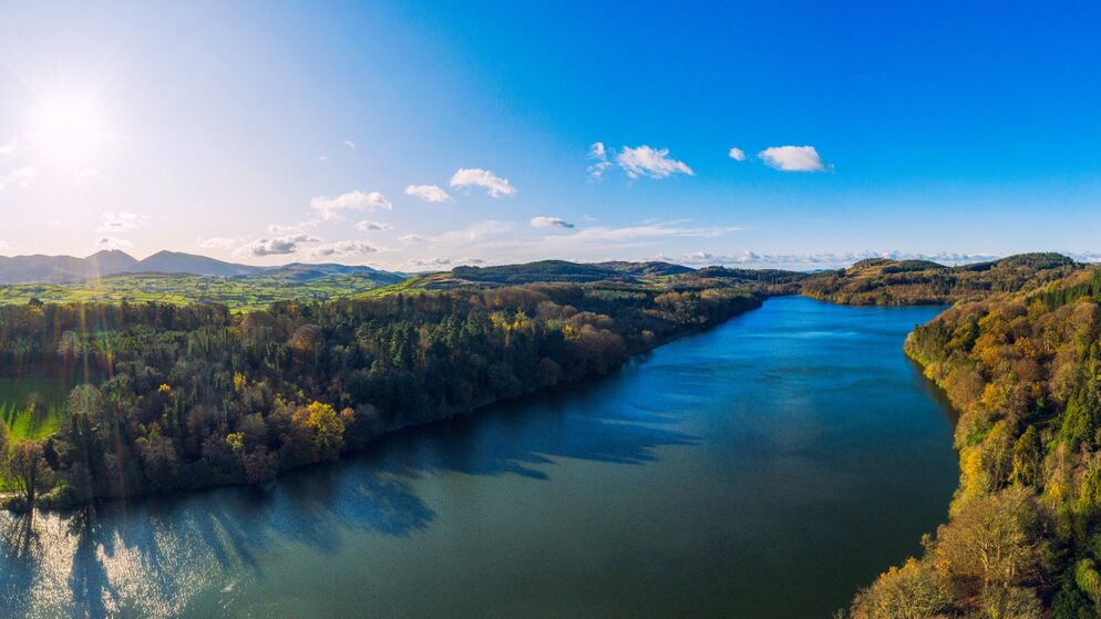 Luftaufnahme von sanften Hügeln, Wäldern und Flusstälern in der Nähe von Belfast an einem sonnigen Tag mit blauem Himmel und Bergen in der Ferne.