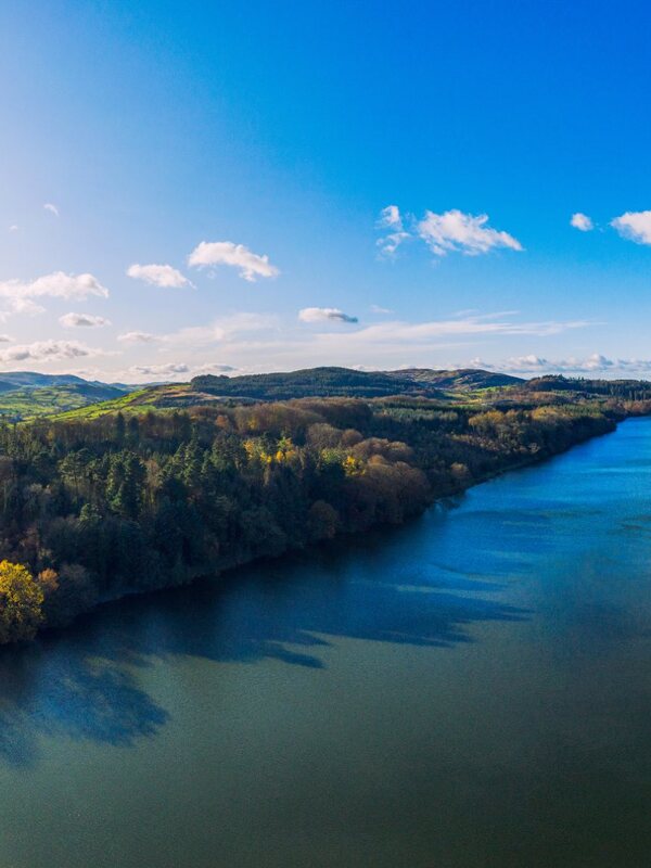 Aerial view of rolling hills, forest, and river valley near Belfast on a sunny day with blue skies and distant mountains.