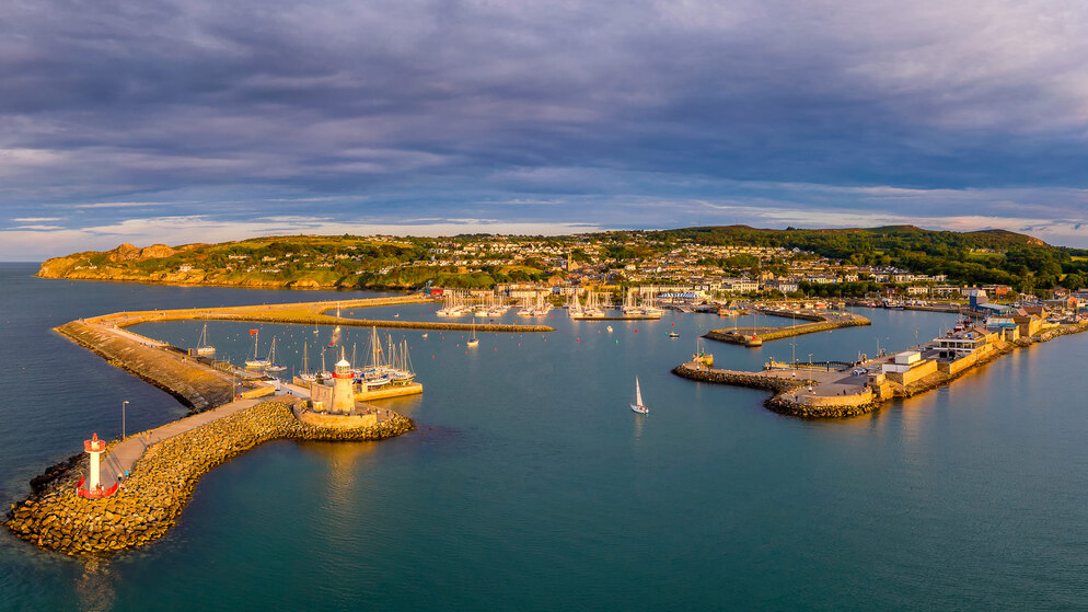 Aerial view of Howth Harbour lined with boats, with golden headlands and village rooftops under dramatic evening skies.