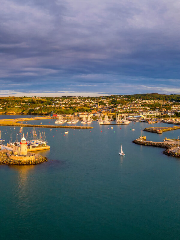 Aerial view of Howth Harbour lined with boats, with golden headlands and village rooftops under dramatic evening skies.