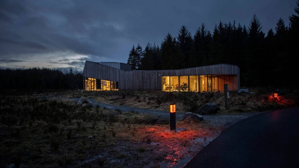 Het moderne, warm verlichte OM Dark Sky Park & Observatory 's nachts in county Tyrone, omgeven door bos onder een donkere hemel.