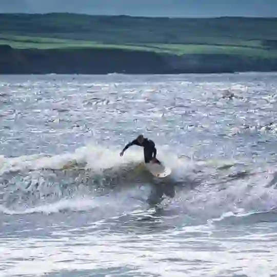 Surfer riding a small breaking wave off the County Clare coast, with rugged green cliffs and shimmering sea in the background.