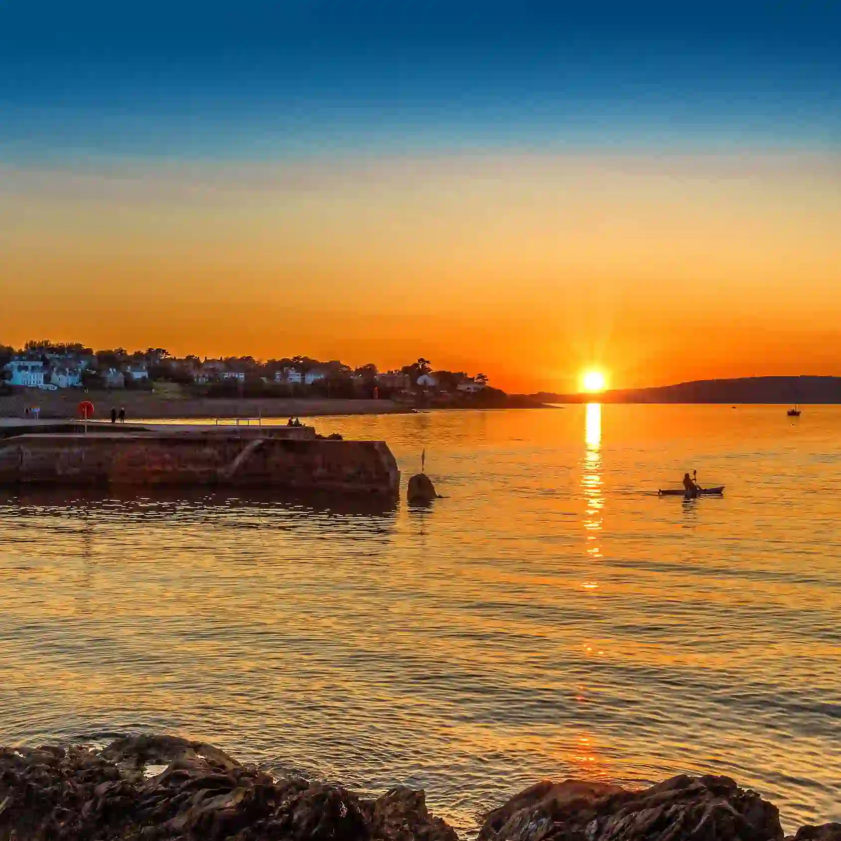 Zonsondergang boven de jachthaven van Bangor, met een gloeiend oranje hemel, kalm water, een steiger en een eenzame kajakker in de baai.