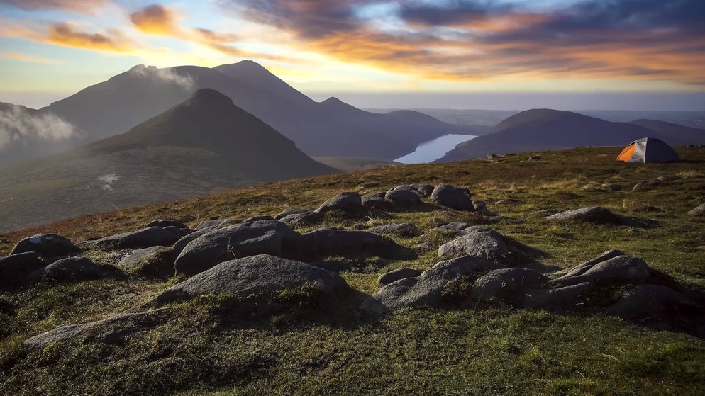 Sunrise over the Mourne Mountains in County Down with rocky peaks, a lakeside valley and a tent overlooking the landscape.