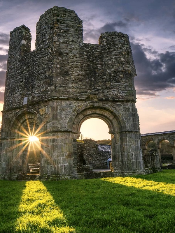 Die Ruinen der Mellifont Abbey in der Grafschaft Louth bei Sonnenuntergang mit antiken Steinbögen, dramatischen Himmeln und langen Schatten über dem Gras.